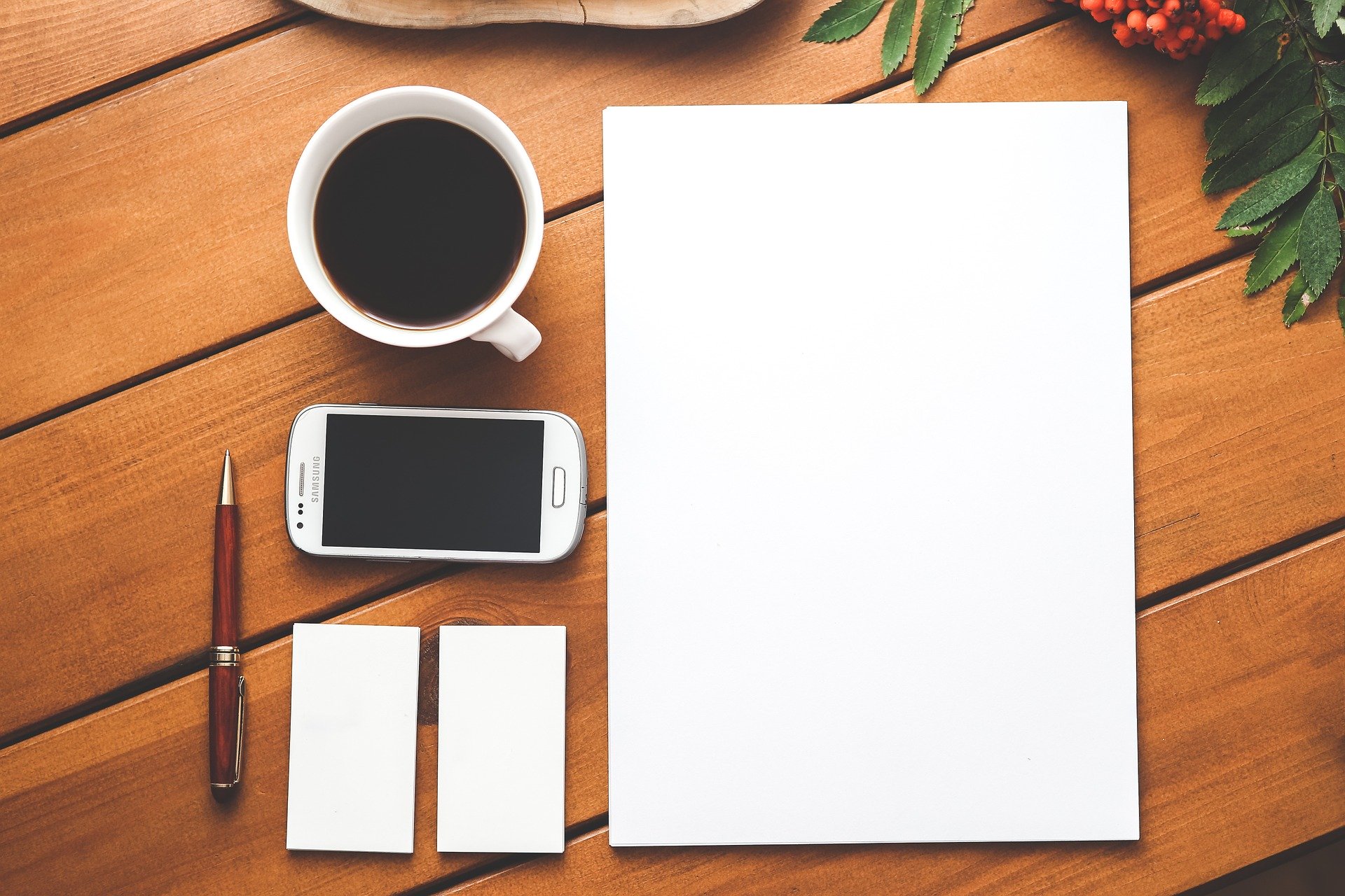 A blank stack of paper sitting next to a cup of coffee, mobile phone, and pen, on a wooden table.